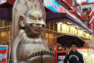 A smiling young person holding an umbrella poses next to a large, seated statue with a mischievous grin in front of a colorful, shop-lined street with Japanese signage.