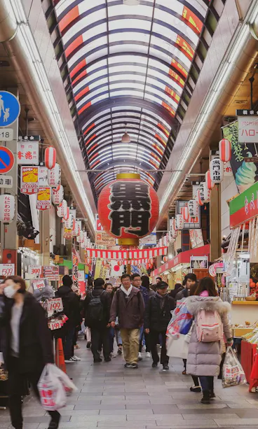 Kuromon Market Indoor Japanese market with people walking and shopping, colorful signs and banners overhead, food stalls and shops line both sides, and a skylight roof lets in natural light.