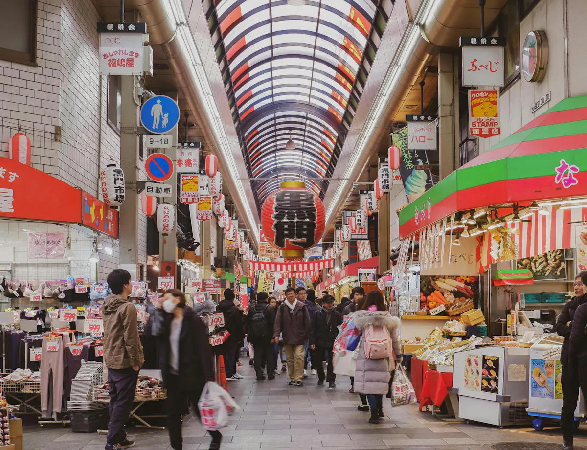 Kuromon Market Indoor Japanese market with people walking and shopping, colorful signs and banners overhead, food stalls and shops line both sides, and a skylight roof lets in natural light.