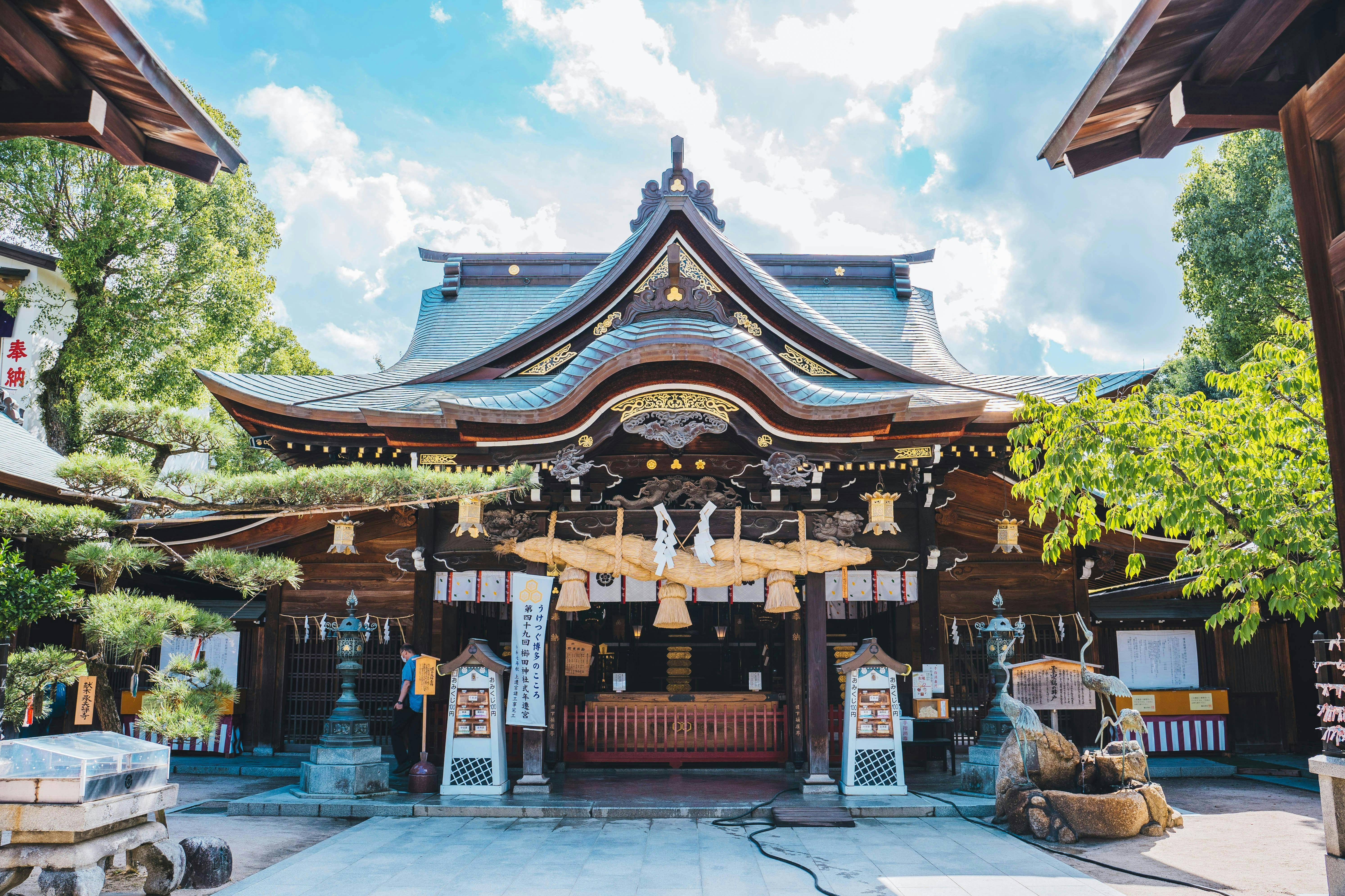 A traditional Japanese Shinto shrine with a curved roof, wooden beams, hanging lanterns, and sacred ropes, surrounded by trees under a partly cloudy sky.