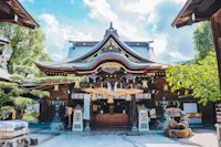A traditional Japanese Shinto shrine with a curved roof, wooden beams, hanging lanterns, and sacred ropes, surrounded by trees under a partly cloudy sky.