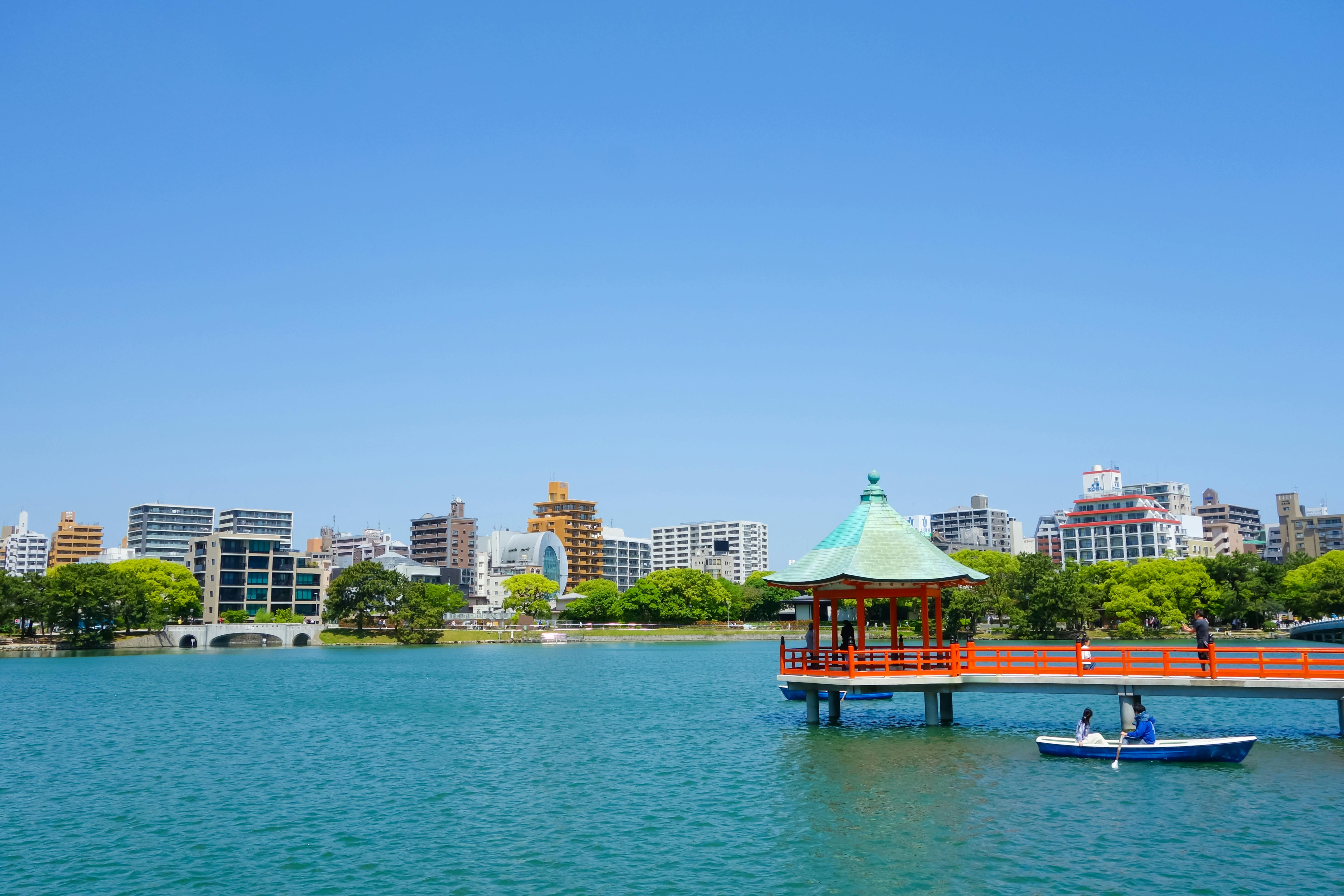 A bright blue lake with a red and green-roofed pavilion on stilts, a small boat with two people on the water, and a city skyline with tall buildings and trees in the background under a clear sky.
