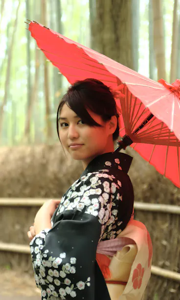 Arashiyama A woman in a black floral kimono holds a red parasol and stands on a path in a bamboo forest, looking back over her shoulder.