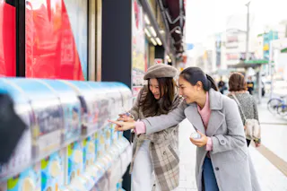 Two young women smiling and excitedly using capsule toy vending machines on a lively city street, with pedestrians and shops in the background.