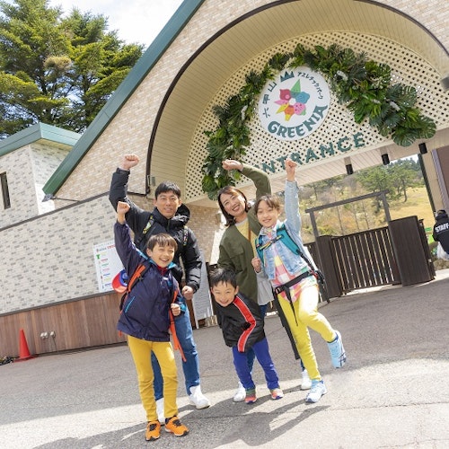Rokkosan GREENIA Athletic Park A smiling family of five poses with raised arms in front of an arched entrance to an outdoor attraction called “Greenia.” The parents and three children wear casual clothes and backpacks, with trees and buildings in the background.