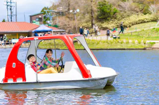 Two children are smiling and waving while riding a red and white pedal boat on a lake, with people and greenery visible in the background on a sunny day.
