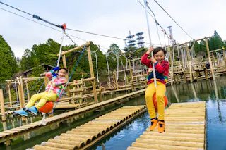 Two children wearing colorful clothes smile as they ride ziplines over a pond, with wooden planks and a ropes course structure in the background, surrounded by trees and tall towers.
