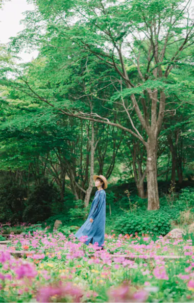 Rokko Alpine Botanical Garden A person in a long blue dress and straw hat walks through a lush garden filled with pink flowers and surrounded by tall green trees.