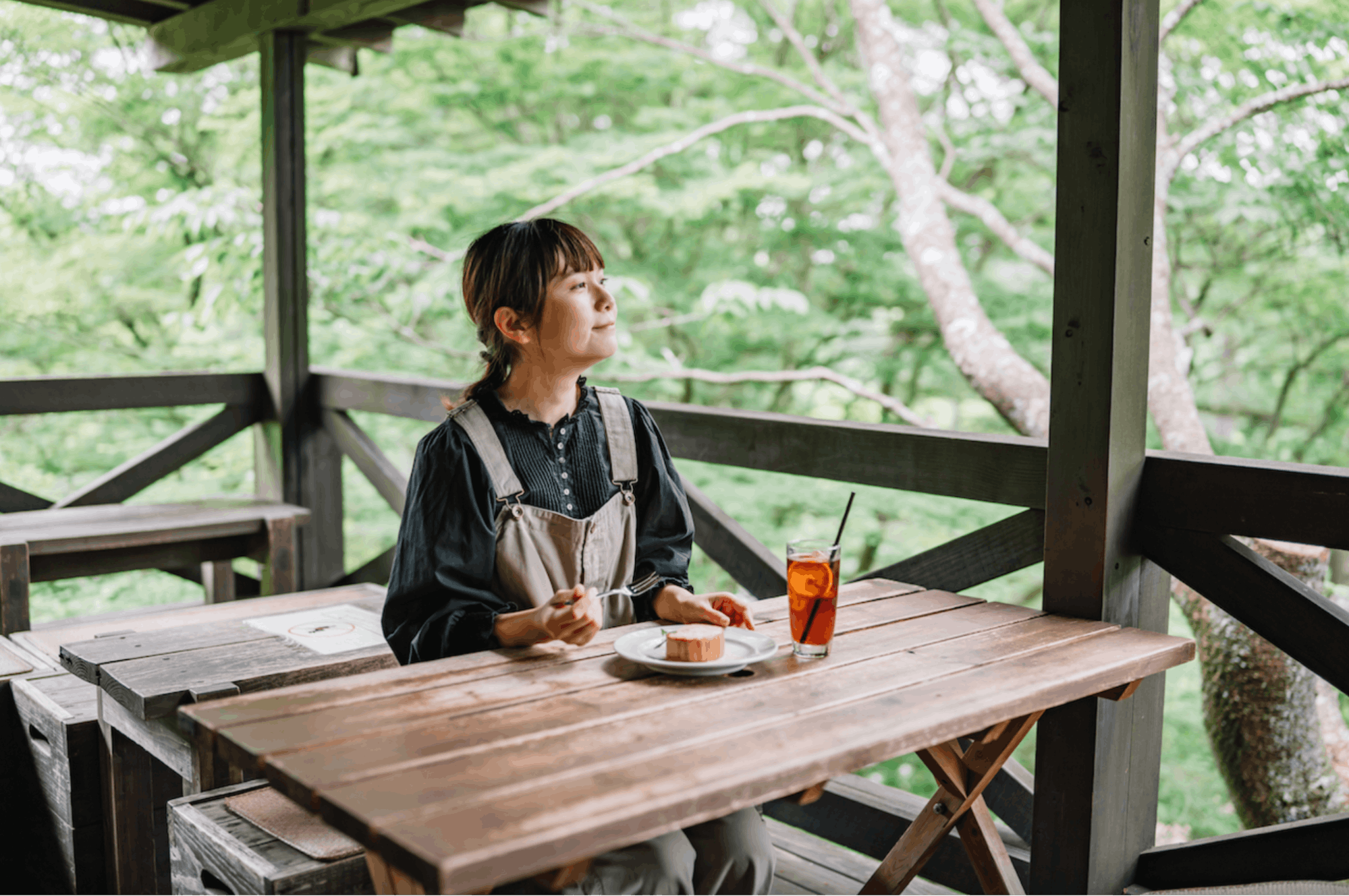 A woman sits alone at a wooden table on an outdoor terrace, enjoying a piece of cake and a glass of iced tea, surrounded by lush green trees.