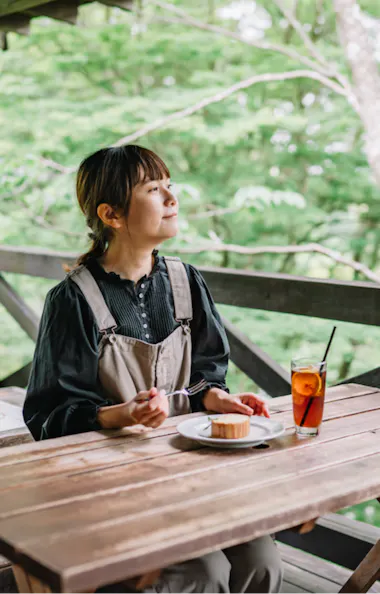 Rokko Alpine Botanical Garden A woman sits alone at a wooden table on an outdoor terrace, enjoying a piece of cake and a glass of iced tea, surrounded by lush green trees.