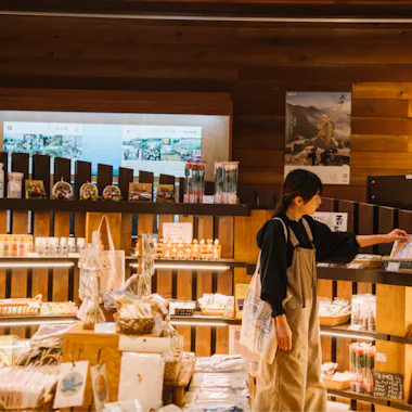 Rokko Alpine Botanical Garden A woman wearing an apron browses shelves stocked with various packaged goods, bags, and baskets in a warmly lit store with wooden walls and display shelves.