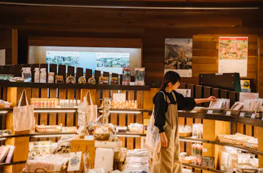 A woman wearing an apron browses shelves stocked with various packaged goods, bags, and baskets in a warmly lit store with wooden walls and display shelves.