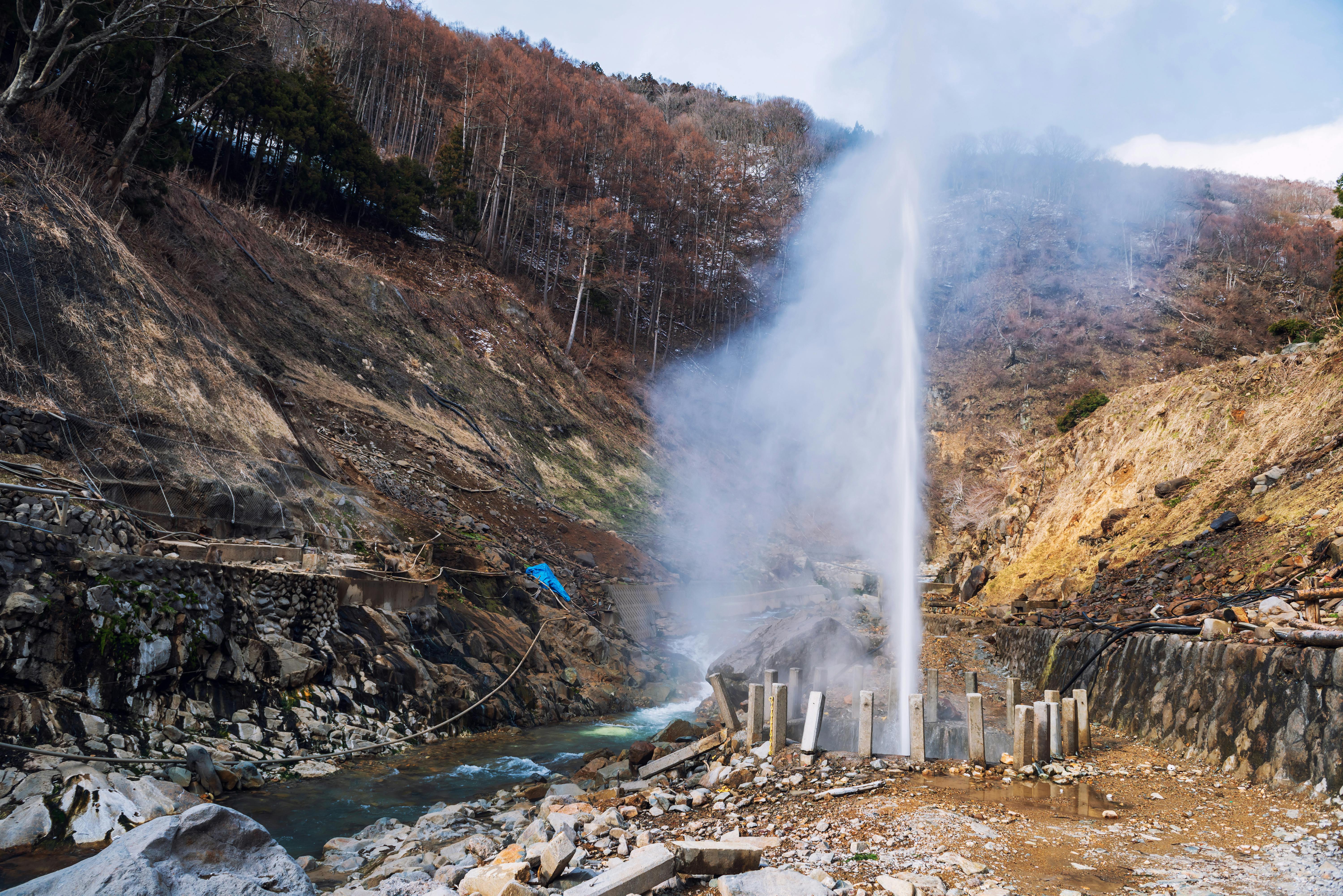 A hot spring geyser shoots a tall column of steam and water into the air amid a rocky, mountainous landscape with sparse vegetation and a stream running nearby.