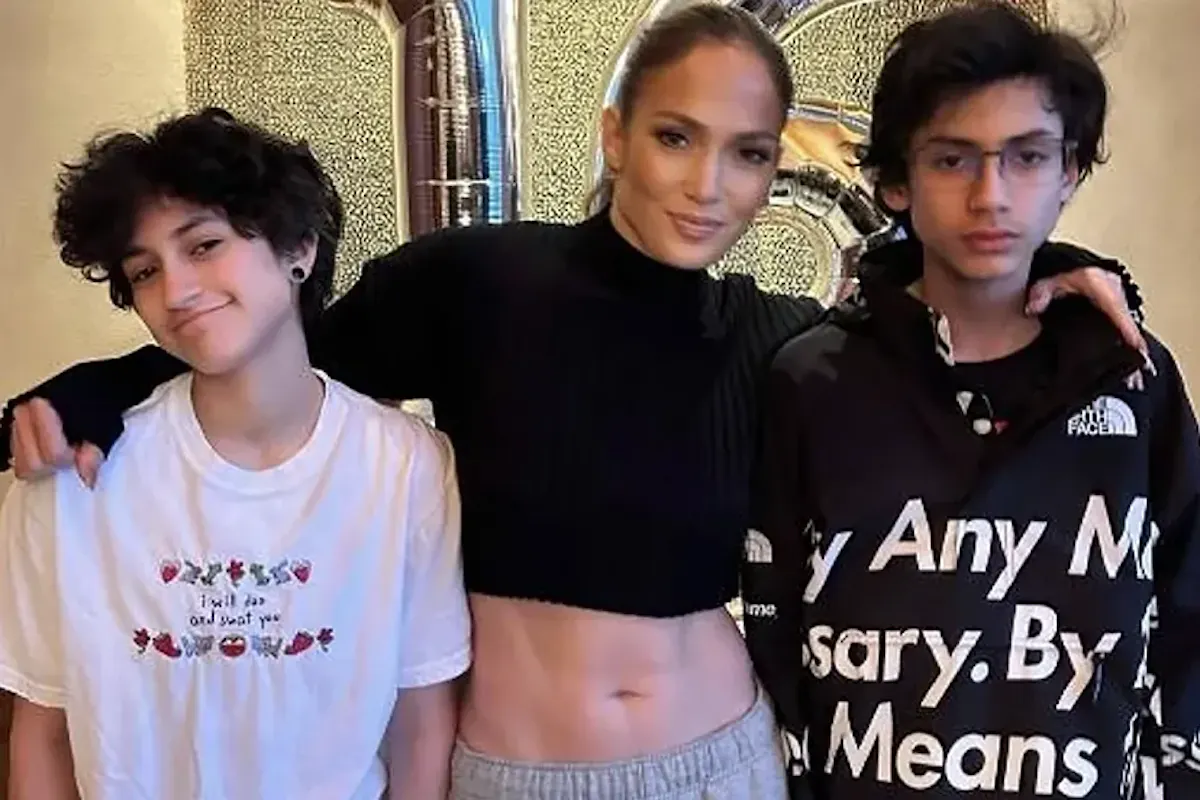 A woman stands in the center with her arms around two teenagers. The woman has her hair pulled back and is wearing a black long-sleeve top, revealing her midriff and gray sweatpants. The teenager on the left has short curly hair, wears a white t-shirt, and smiles. The teenager on the right has dark hair, glasses, and wears a black graphic hoodie. Silver number balloons are in the background.