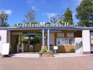 The entrance to Garden Museum Hiei, featuring a green "ENTRANCE" sign, information boards, ticket windows, and surrounding trees under a clear blue sky.