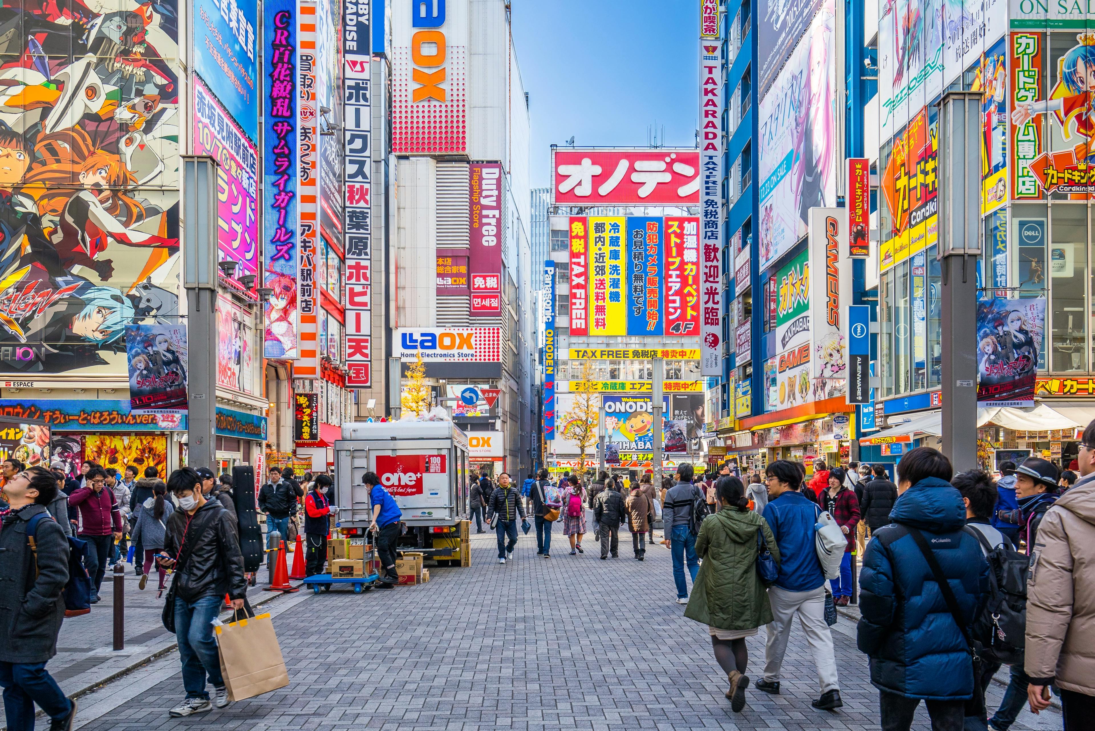 A busy street in Akihabara, Tokyo, filled with people walking among colorful buildings covered in bright advertisements, anime posters, and electronics store signs. The atmosphere is lively and urban.