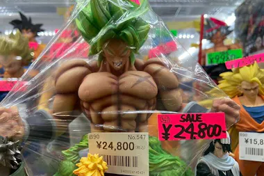 A muscular anime action figure with green hair is wrapped in plastic and displayed for sale. Price tags show ¥24,800, with a sale sign in red. Other action figures are visible in the background.