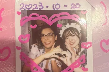 A decorated instant photo shows two smiling people, one wearing glasses and the other in a maid headband, making a heart shape with their hands. The border is adorned with pink hearts and the date "2023-10-20" is written on top.