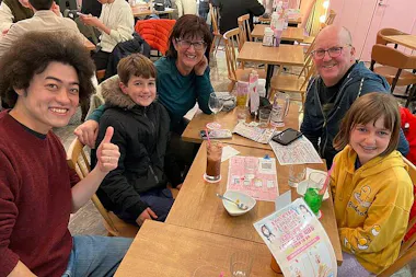 Five people, including two children and three adults, smile and pose for a photo at a café table with drinks, menus, and empty dishes. One man gives a thumbs up. The setting is casual and cheerful.
