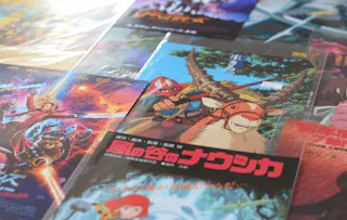 Several colorful Japanese anime and movie posters are spread out on a surface, including a prominent "Nausicaä of the Valley of the Wind" cover with a girl riding a creature, surrounded by other illustrated posters.