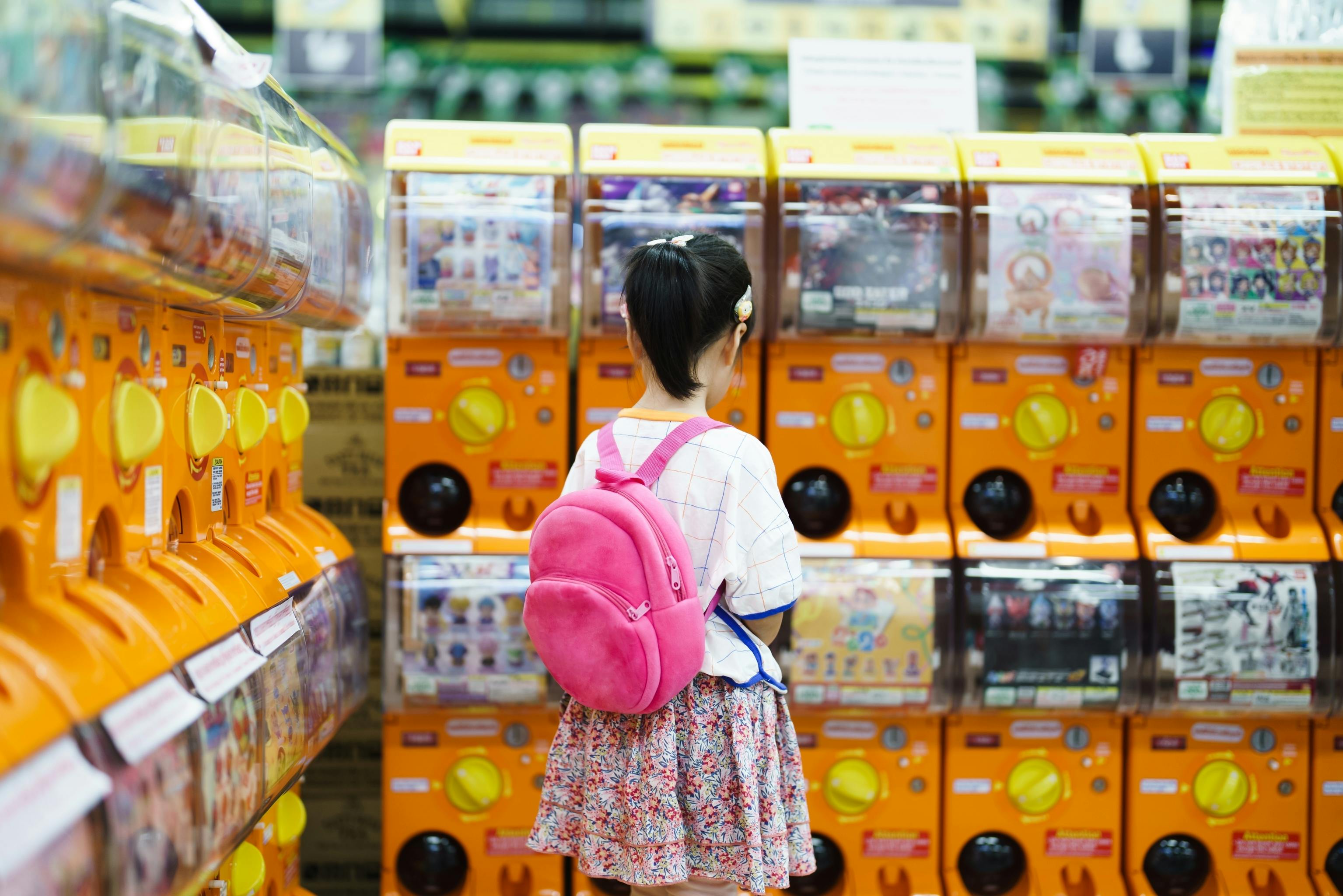 A young girl with a pink backpack stands in front of a row of orange capsule toy vending machines in a brightly lit store.