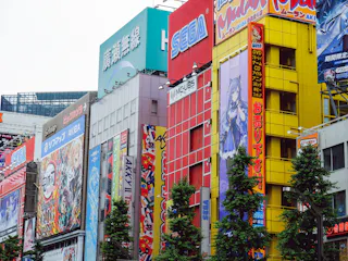 Colorful buildings in Akihabara, Tokyo, covered with large anime, manga, and game advertisements, including a bright yellow building with a character poster and a blue SEGA sign. Green trees line the street in front.