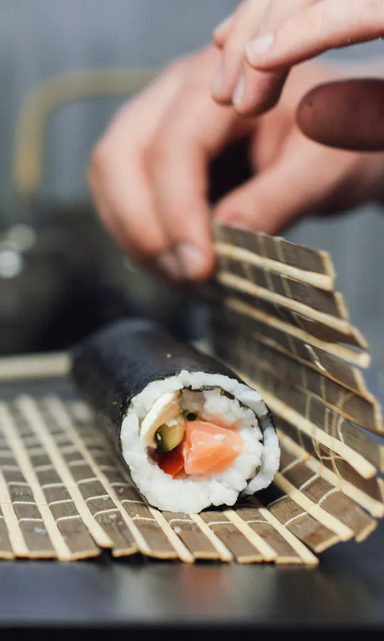 Making Sushi Hands rolling sushi with seaweed, rice, and fish on a bamboo mat, with a blurred plate of sushi and a teapot in the background.