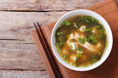 A bowl of miso soup with tofu cubes, chopped green onions, and seaweed on a bamboo mat, with a pair of wooden chopsticks beside it on a rustic wooden table.