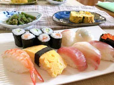 A plate of assorted sushi including nigiri with shrimp, egg, and fish, as well as maki rolls with cucumber and salmon. In the background are side dishes of asparagus and tamagoyaki (Japanese omelette).