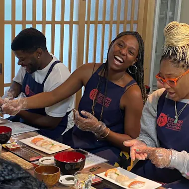 Sushi Class Tokyo Three people wearing aprons and plastic gloves sit at a table making sushi. The woman in the middle is laughing, while the others focus on shaping rice and fish. Sushi ingredients and dishes are arranged in front of them.