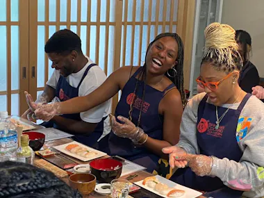 Three people wearing aprons and plastic gloves sit at a table making sushi. The woman in the middle is laughing, while the others focus on shaping rice and fish. Sushi ingredients and dishes are arranged in front of them.