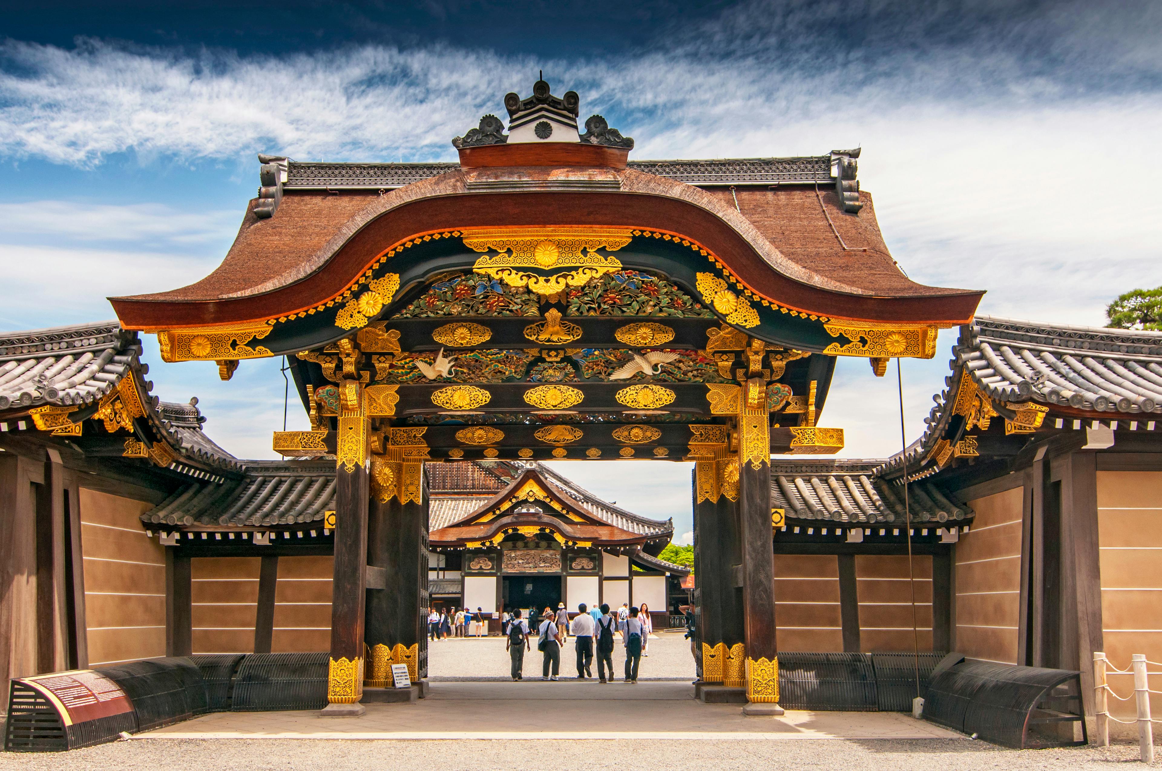 A group of people walk through an ornate wooden gate with intricate gold decorations at Nijo Castle in Kyoto, Japan, under a blue sky with clouds. Traditional buildings are visible in the background.