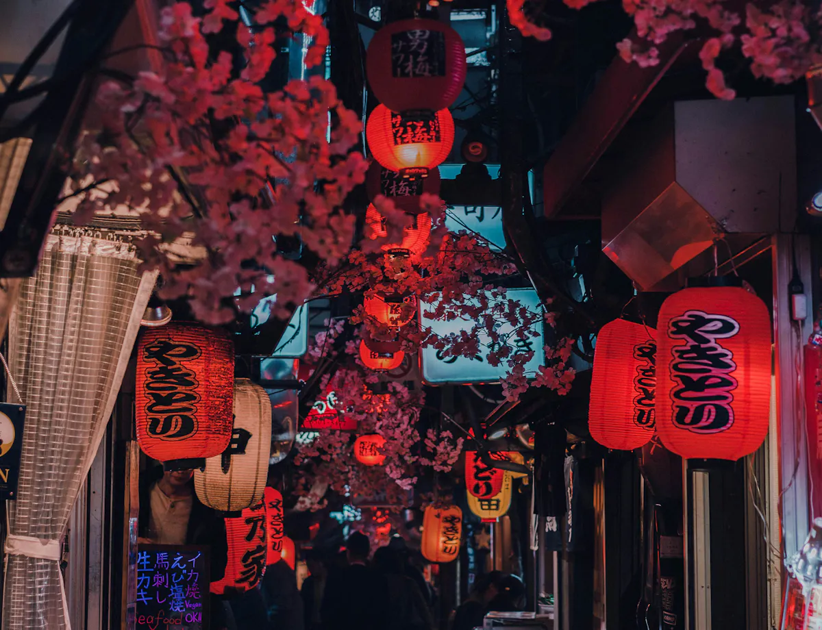 Omoide Yokocho A narrow alleyway in Japan, lit by numerous red paper lanterns with Japanese writing, adorned with pink cherry blossom decorations and lined with small shops and restaurant signs. People can be seen walking in the distance.