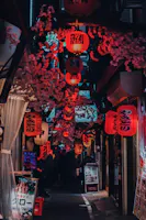 A narrow alleyway in Japan, lit by numerous red paper lanterns with Japanese writing, adorned with pink cherry blossom decorations and lined with small shops and restaurant signs. People can be seen walking in the distance.