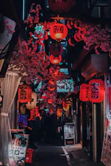 A narrow alleyway in Japan, lit by numerous red paper lanterns with Japanese writing, adorned with pink cherry blossom decorations and lined with small shops and restaurant signs. People can be seen walking in the distance.