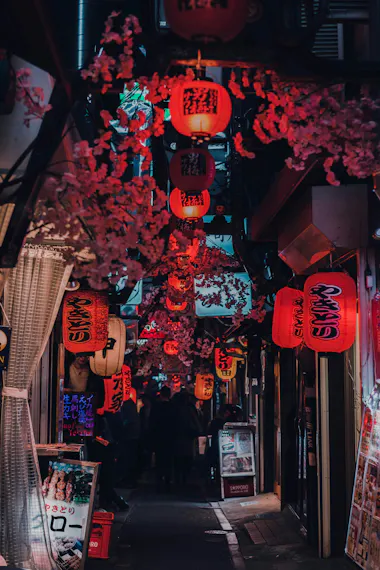 A narrow alleyway in Japan, lit by numerous red paper lanterns with Japanese writing, adorned with pink cherry blossom decorations and lined with small shops and restaurant signs. People can be seen walking in the distance.