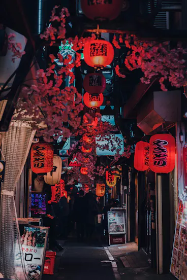 A narrow alleyway in Japan, lit by numerous red paper lanterns with Japanese writing, adorned with pink cherry blossom decorations and lined with small shops and restaurant signs. People can be seen walking in the distance.