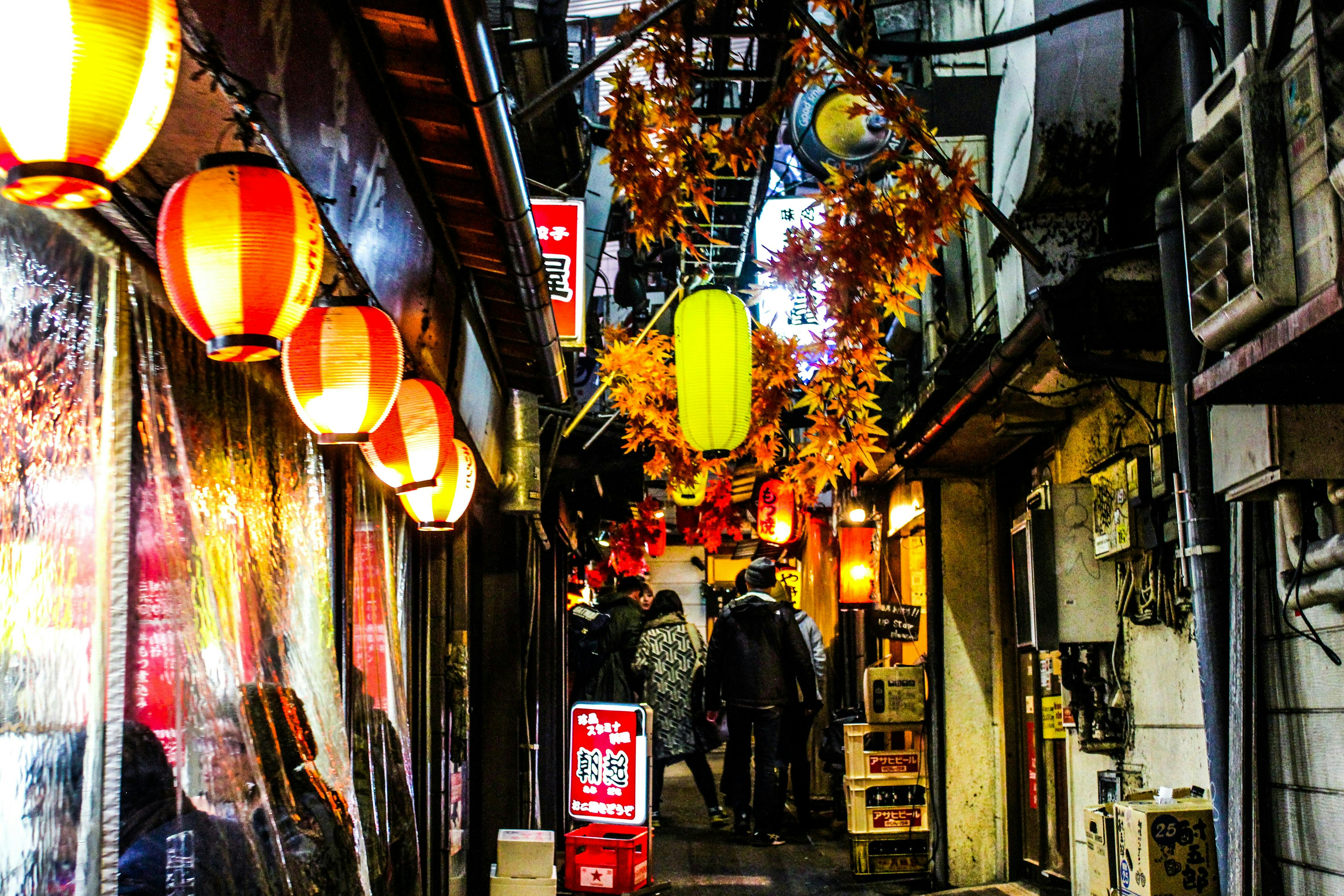 A narrow alleyway in Japan decorated with red and yellow lanterns and artificial autumn leaves, with people walking and small shops lining both sides. The scene is vibrant and lively, evoking a festive atmosphere.