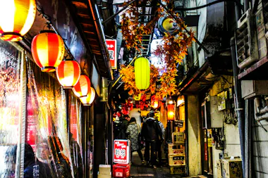 A narrow alleyway in Japan decorated with red and yellow lanterns and artificial autumn leaves, with people walking and small shops lining both sides. The scene is vibrant and lively, evoking a festive atmosphere.