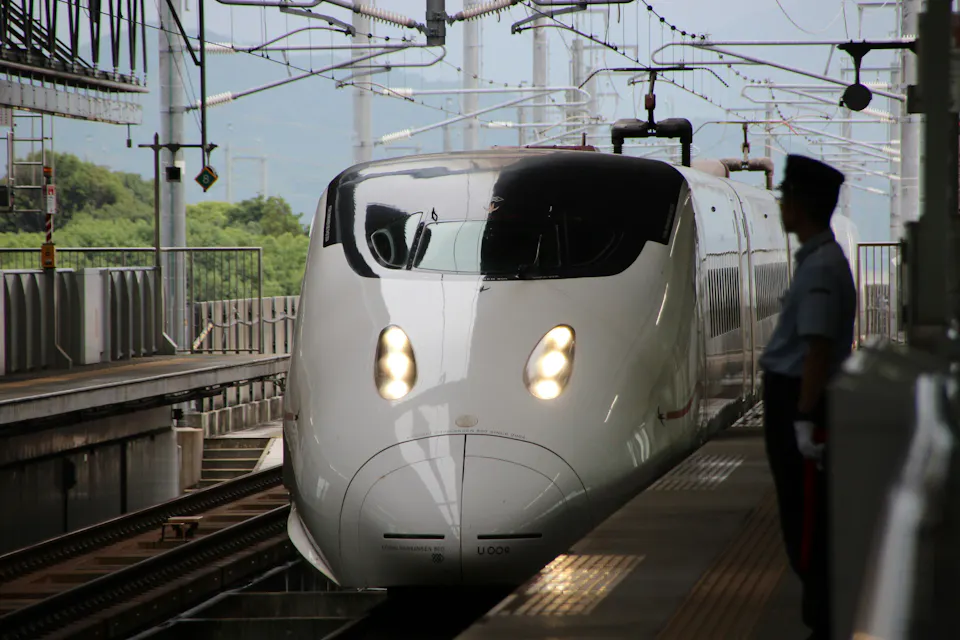A white bullet train approaches a platform under a canopy at a station. The train's headlights are on, and it appears to be slowing down or stopping. A security guard in a light blue uniform and cap stands by the side, monitoring the arrival.