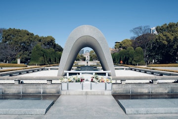 A peaceful scene of the Hiroshima Peace Memorial Park, featuring the Memorial Cenotaph with a stone arch and flowers, surrounded by neatly trimmed trees and a clear blue sky in the background.