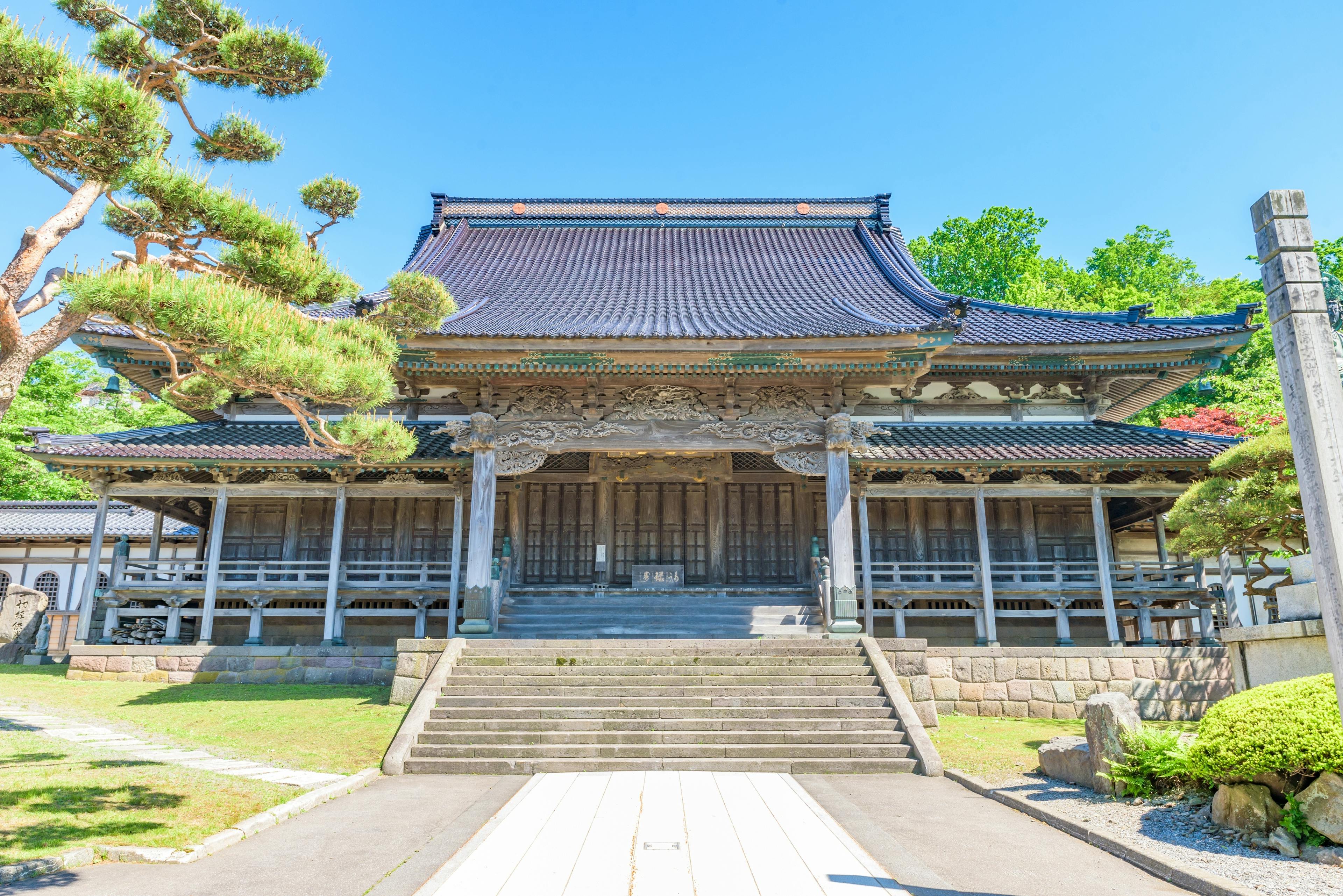 Traditional Japanese temple with wooden architecture and ornate roof, surrounded by greenery and a bright blue sky, viewed from the front with steps leading up to the entrance.
