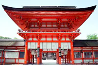 Bright red traditional Japanese shrine gate with white accents and intricate wooden details, standing against a clear sky. The entrance leads to a peaceful temple courtyard visible through the gate.