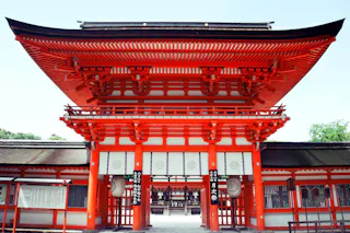 Bright red traditional Japanese shrine gate with white accents and intricate wooden details, standing against a clear sky. The entrance leads to a peaceful temple courtyard visible through the gate.