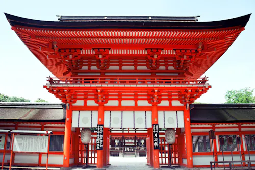 Bright red traditional Japanese shrine gate with white accents and intricate wooden details, standing against a clear sky. The entrance leads to a peaceful temple courtyard visible through the gate.