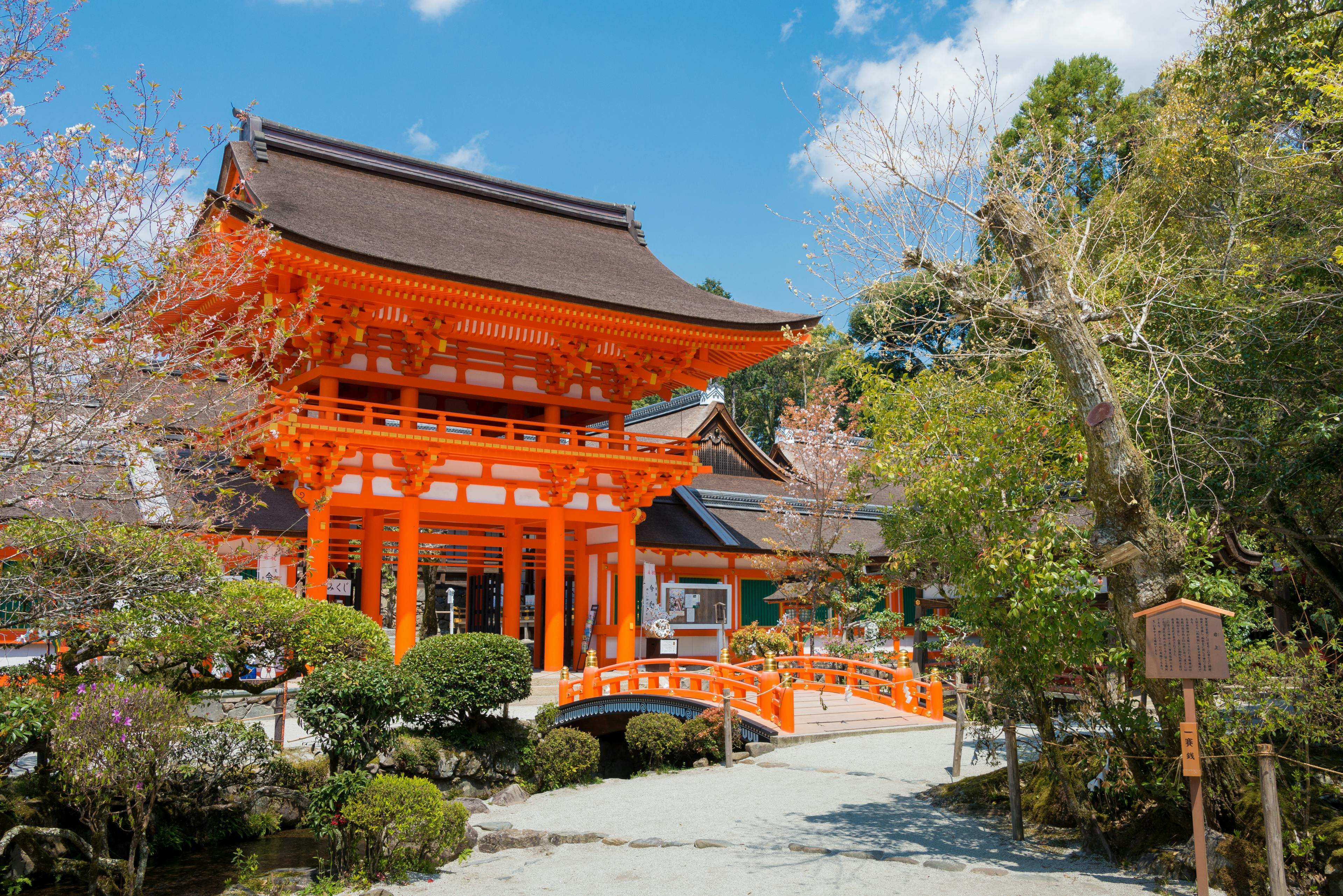 A bright orange traditional Japanese shrine gate stands near a small arched bridge, surrounded by trees, shrubs, and a clear blue sky.