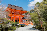 A bright orange traditional Japanese shrine gate stands near a small arched bridge, surrounded by trees, shrubs, and a clear blue sky.