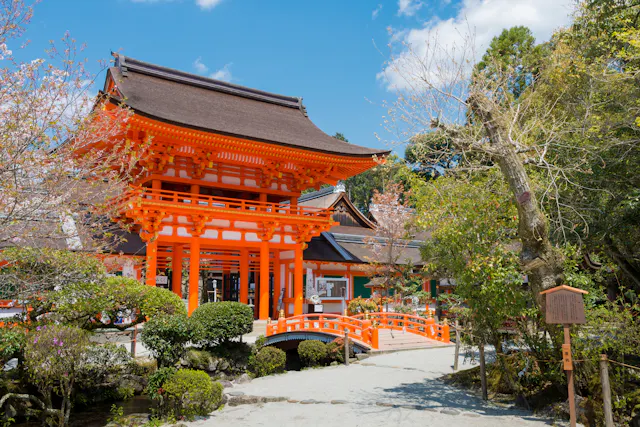 A bright orange traditional Japanese shrine gate stands near a small arched bridge, surrounded by trees, shrubs, and a clear blue sky.