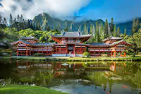 A traditional red Japanese temple with ornate rooftops stands beside a calm pond, surrounded by lush greenery and tall mountains partly covered by clouds in the background.