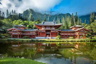 A traditional red Japanese temple with ornate rooftops stands beside a calm pond, surrounded by lush greenery and tall mountains partly covered by clouds in the background.